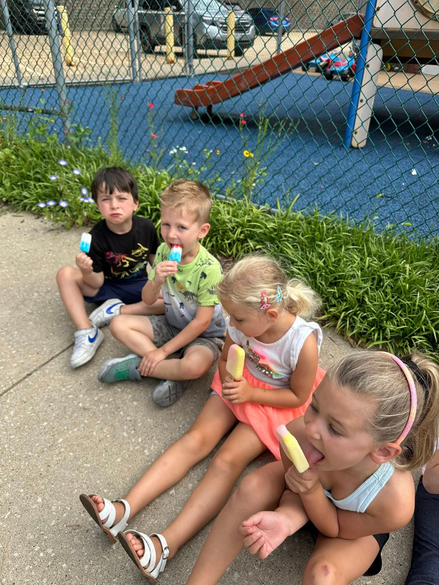 Four preschoolers eating popsicles beside the playground fence