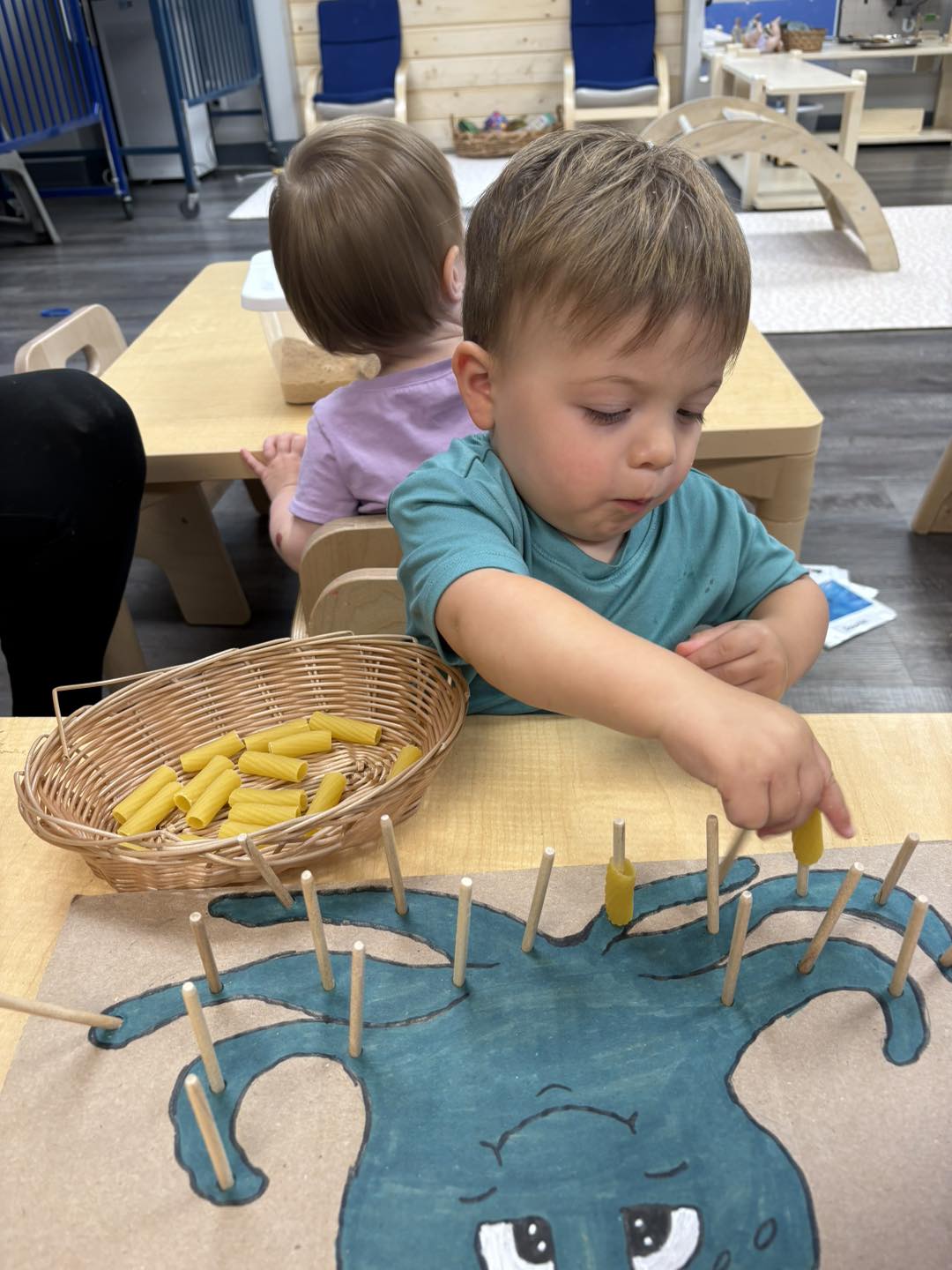 A toddler threading dry pasta onto wooden pegs at a low table