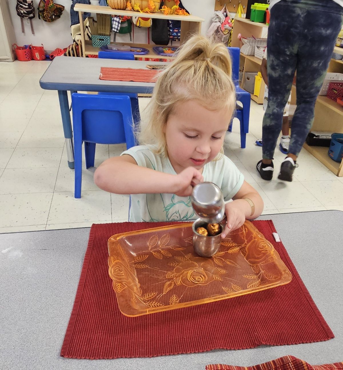 A preschool girl carefully pouring from a small metal pitcher on a work mat