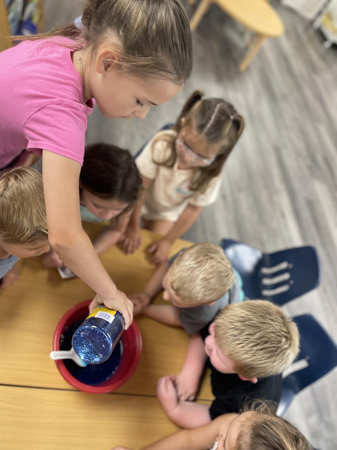 Children gathered around a bowl as one pours blue glitter into water