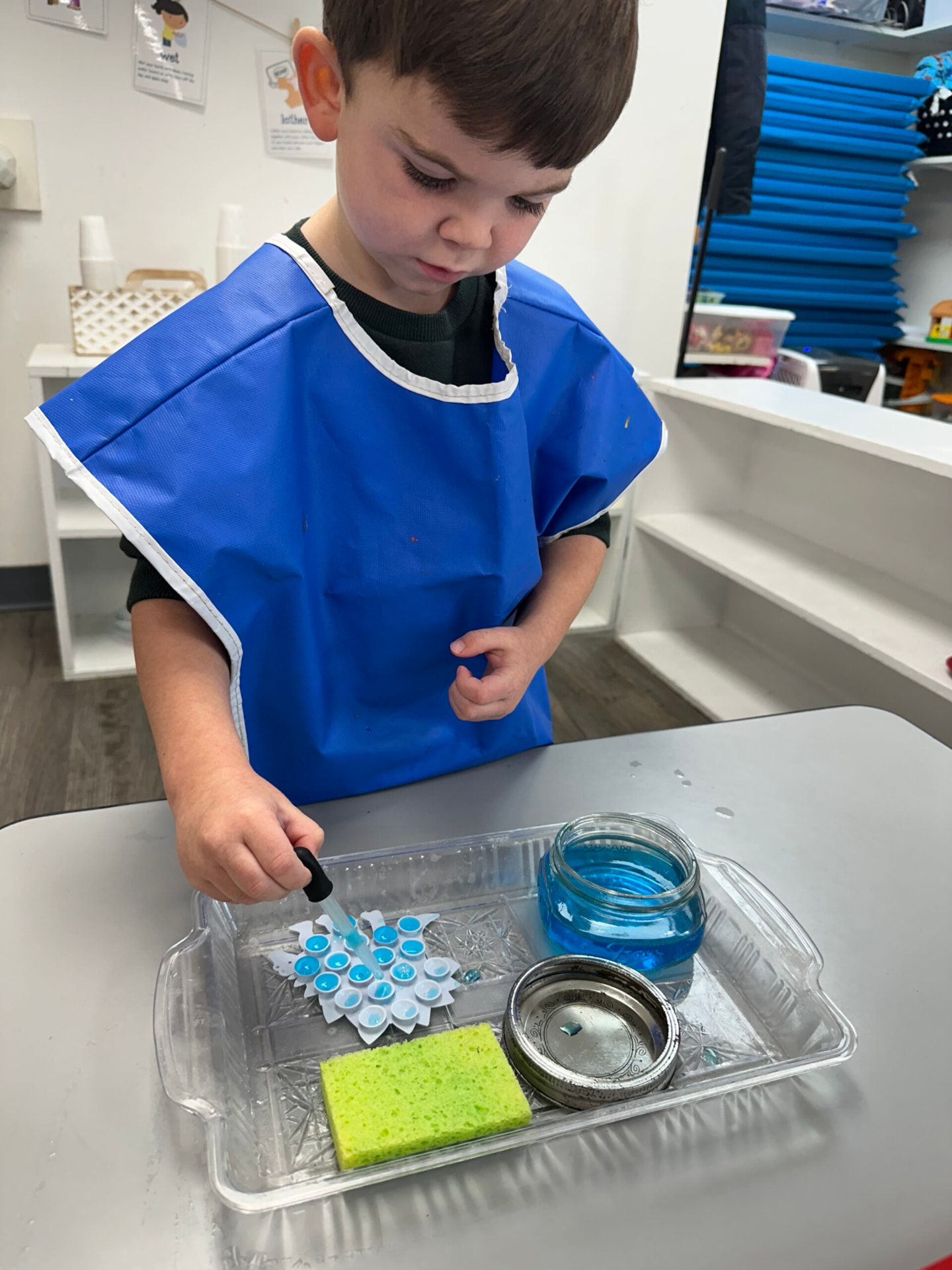 A boy in a blue smock using a dropper to fill a snowflake tray with blue water