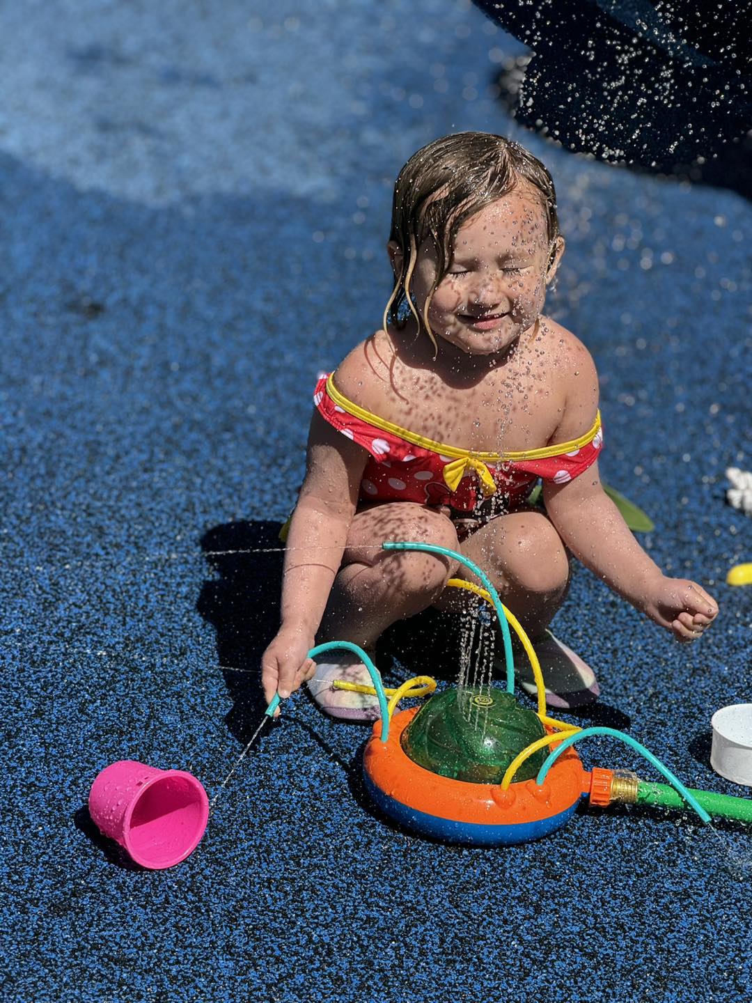 A toddler crouched beside a sprinkler toy, laughing in the spray