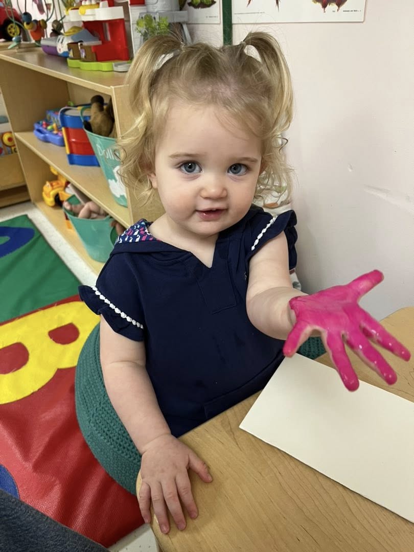 A toddler showing off a pink painted hand at a classroom table