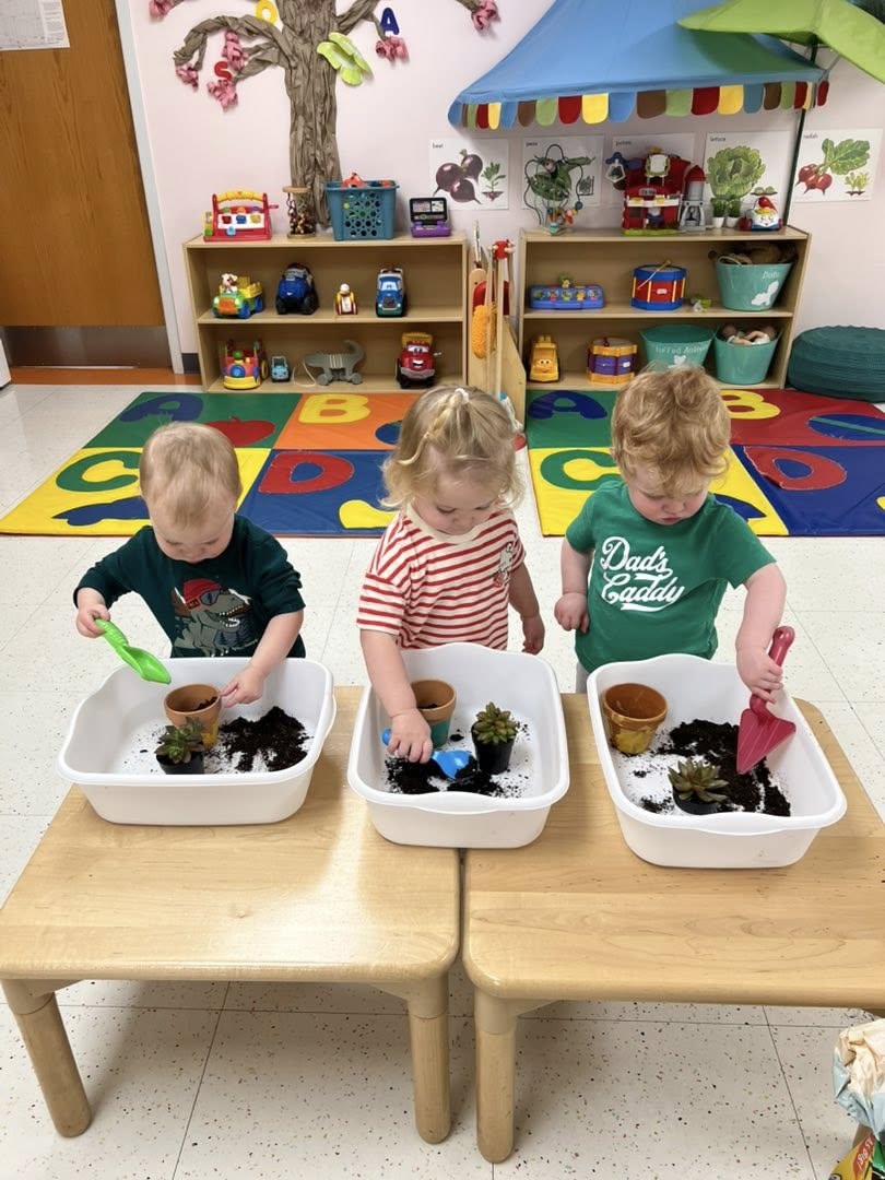 Three toddlers scooping soil into small pots at low tables