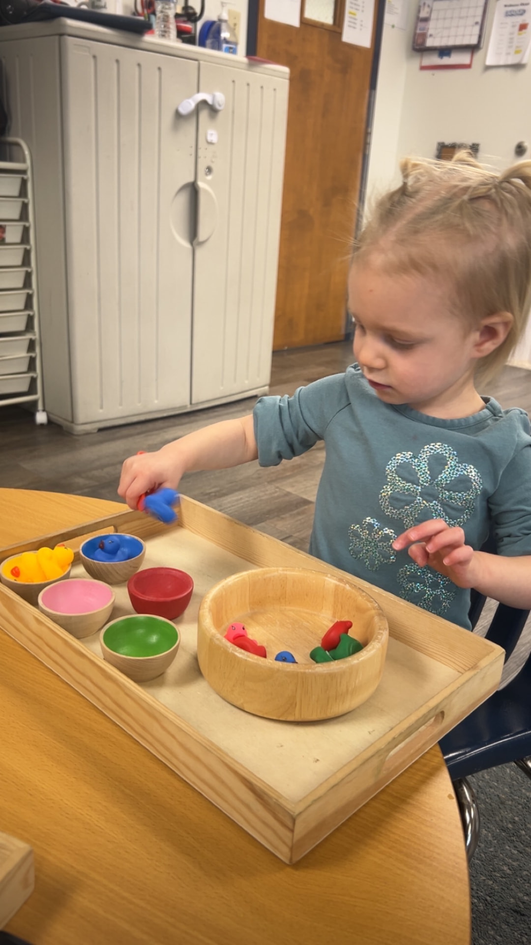 A toddler sorting small rubber ducks into colored bowls on a wooden tray