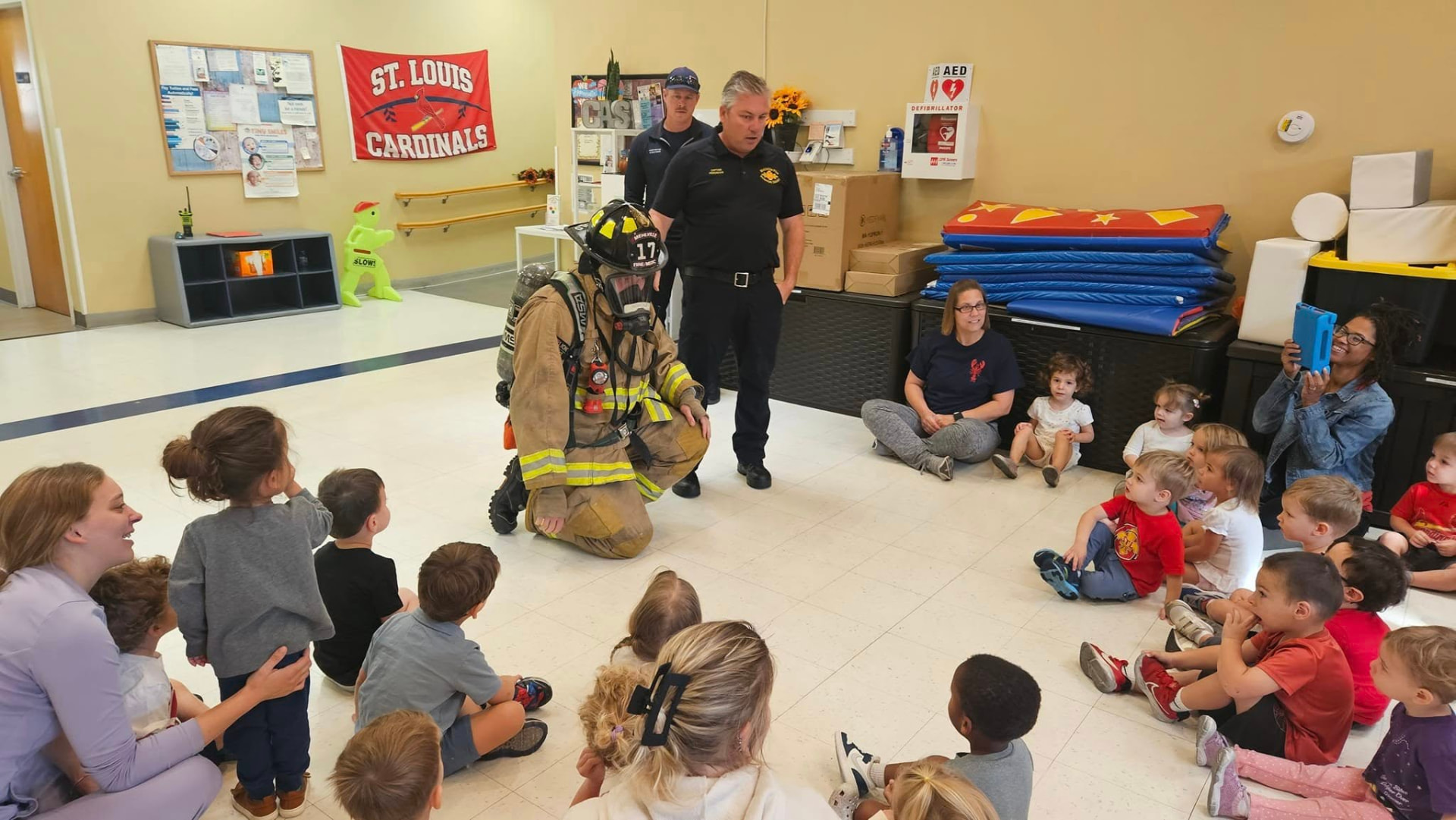 Children seated in a circle listening to firefighters in full gear