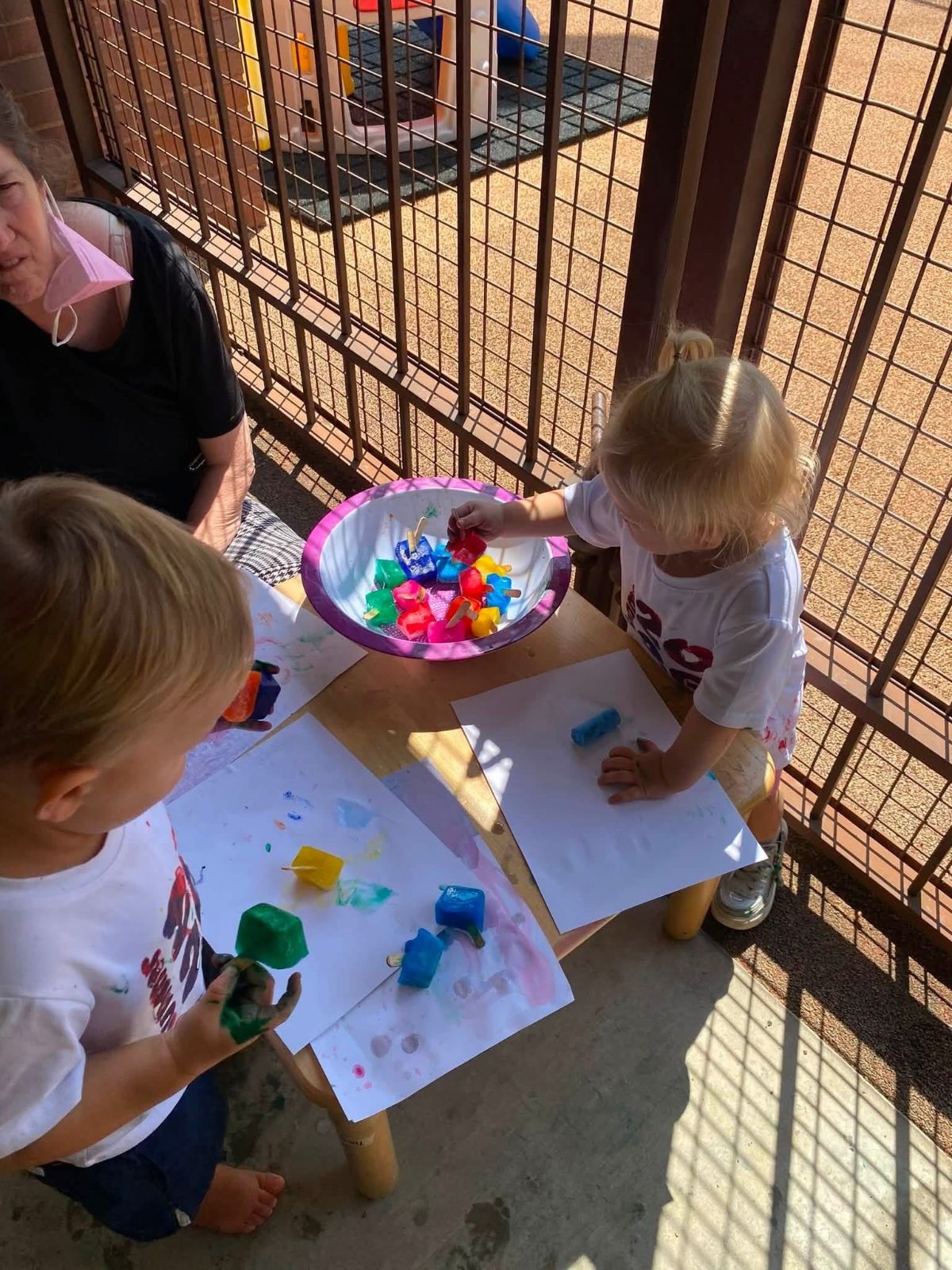Toddlers painting with colored ice cubes on a shaded porch